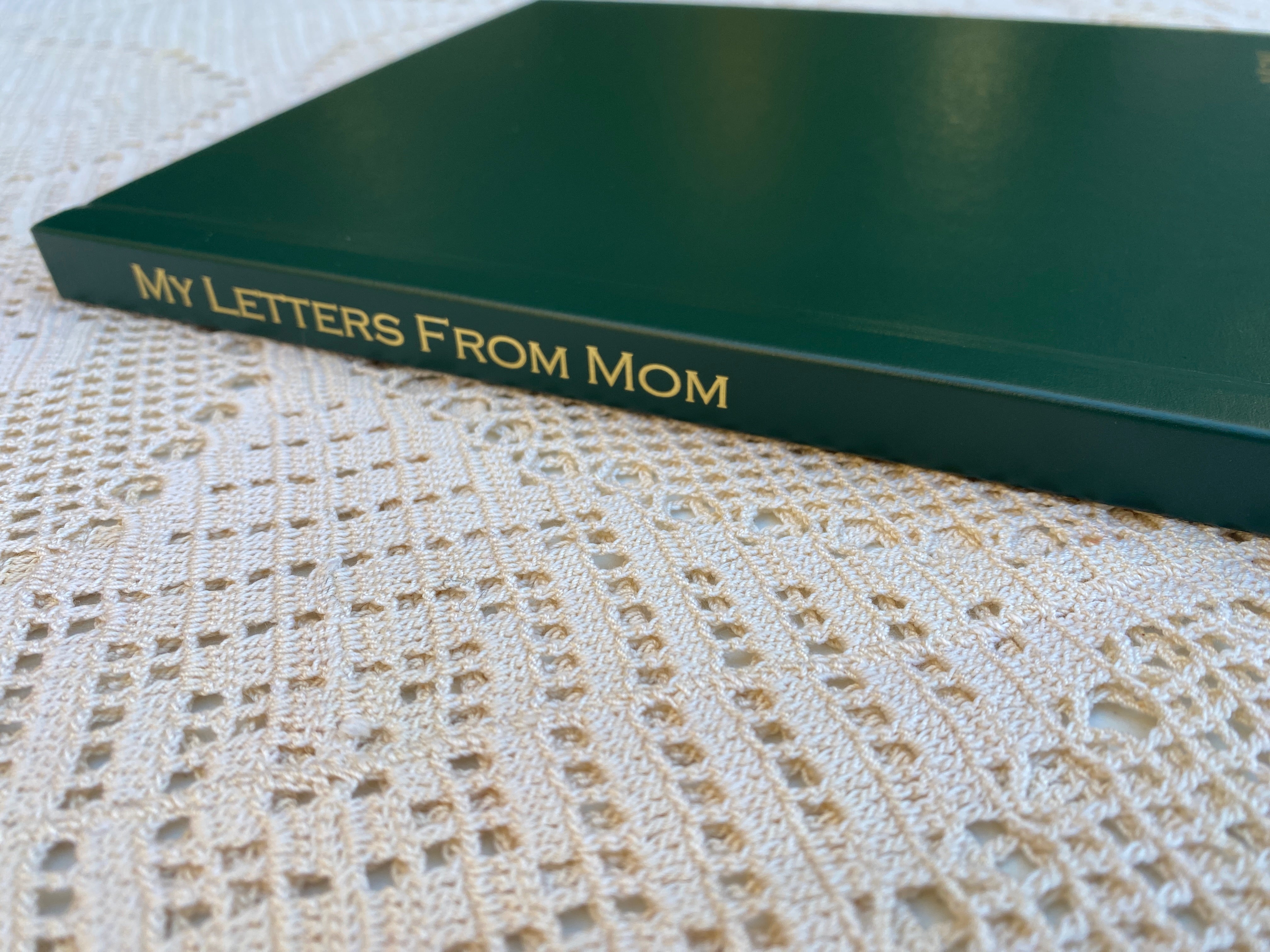 Green custom hardcover book with gold foil spine title “My Letters From Mom” on a cream lace tablecloth, a letter keepsake book by 4everBound